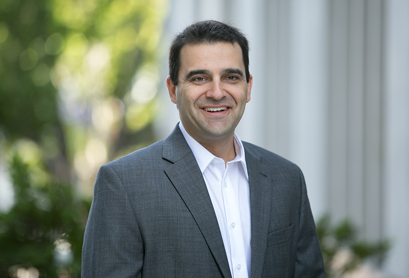 Man in a dark gray jacket and white collared shirt standing in front of a building