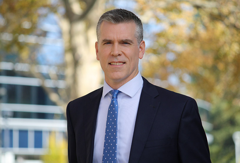 Photo of a men with a blue tie and a black jacket standing in front of a building.
